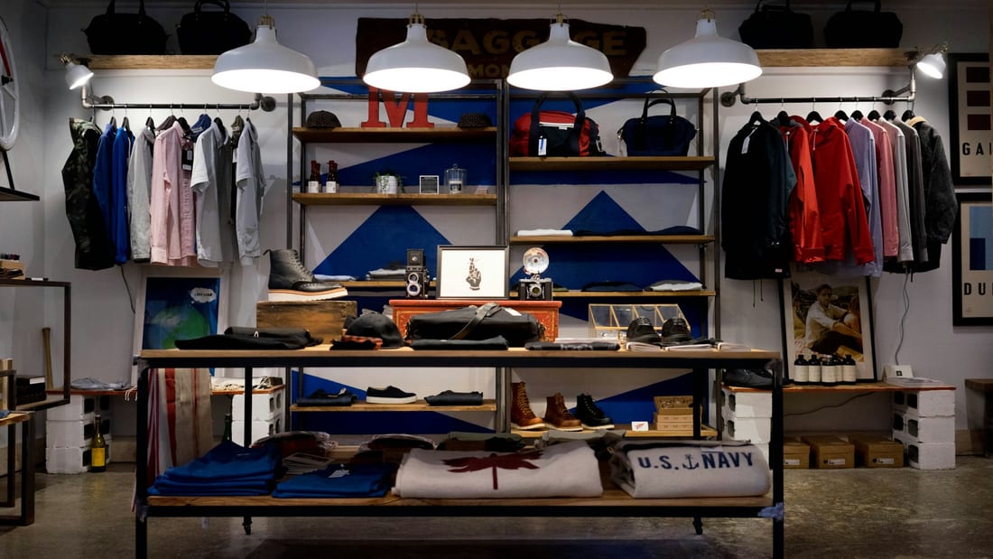 The interior of a menswear shop: rails of shirts and jackets, a central display table of folded knitwear and boots, under warm pendant lighting.